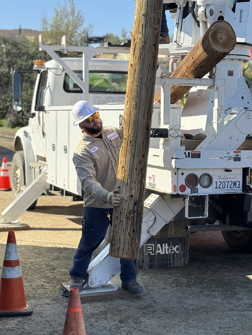 An Anza Electric Coop utility worker wearing a white construction helmet helps guide a utility pole from off a big utility truck
