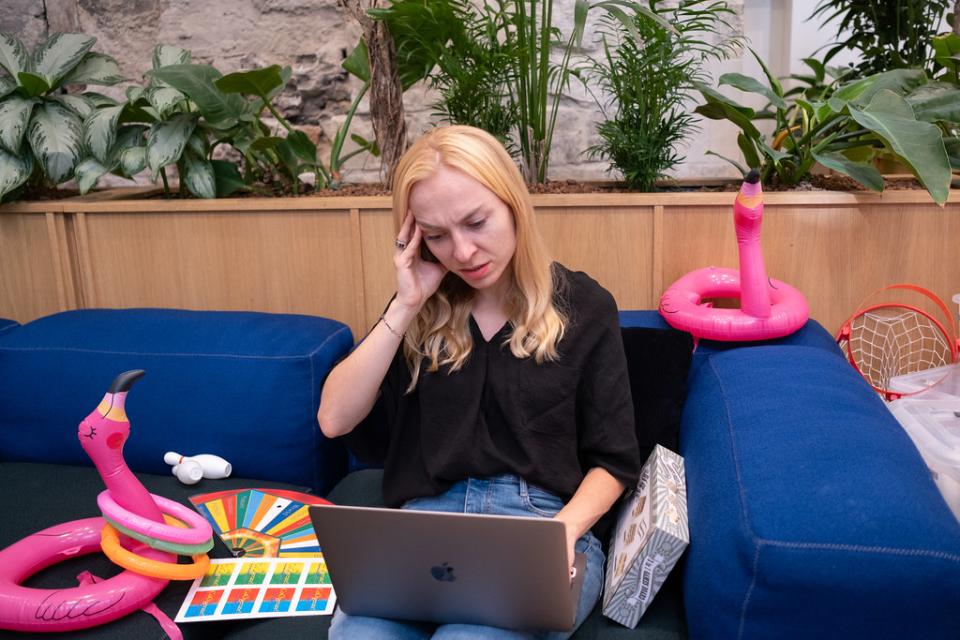 A mom is distressed with her hand on her temple looking at the laptop on her lap, seated next to childrens toys